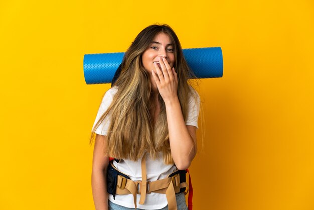 Mujer joven alpinista con una mochila grande en amarillo feliz y sonriente cubriendo la boca con la mano