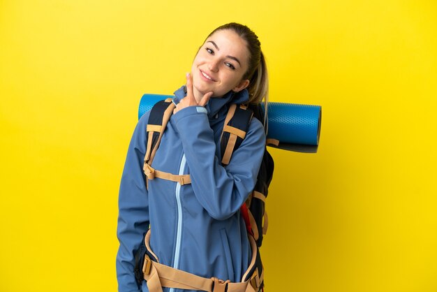 Mujer joven alpinista con una mochila grande sobre fondo amarillo aislado feliz y sonriente