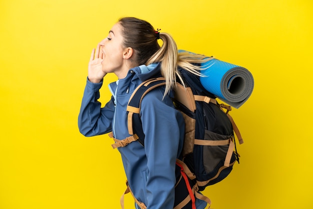 Mujer joven alpinista con una mochila grande sobre fondo amarillo aislado gritando con la boca abierta hacia el lado