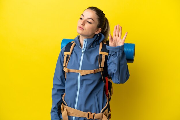 Mujer joven alpinista con una mochila grande sobre fondo amarillo aislado haciendo gesto de parada y decepcionado