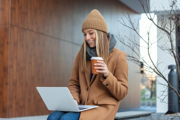 Mujer joven con una amplia sonrisa sentada en el banco mira la computadora portátil con café en las manos en el administrador de la calle de la ciudad que no es de oficina se dedica a un trabajo adicional en la computadora portátil