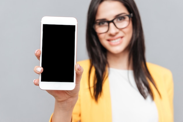 Mujer joven con anteojos y vestida con chaqueta amarilla que muestra la pantalla del teléfono al frente sobre una superficie gris. Mira al frente. Concéntrese en el teléfono.