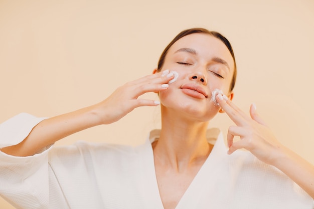 Mujer joven aplicando crema cosmética blanca en el rostro