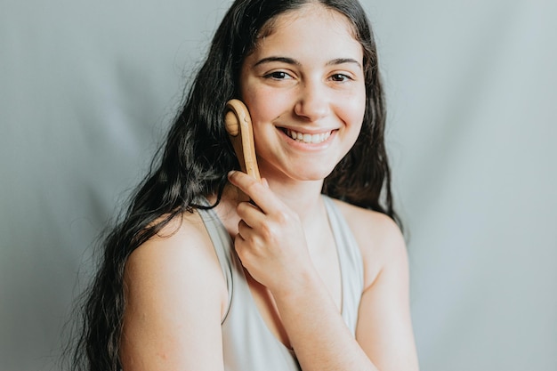 Mujer joven aplicando masaje facial con rollo de madera Alegre día de autocuidado Aplicar presión para mejorar la circulación Nuevos métodos de cuidado de la piel y rituales diariosRetrato en estudio con fondo gris