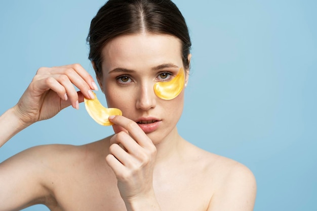 Mujer joven aplicando parches dorados debajo de los ojos mirándose en el espejo aislado de fondo azul