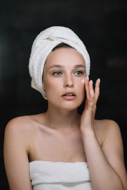 Mujer joven aplicar crema facial mirándose en el espejo hermosa mujer sana envuelta en toallas poner crema hidratante de día nutritiva en piel suave hidratada