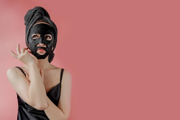 Mujer joven aplicar máscara facial de tela cosmética negra sobre fondo rosa Máscara de peeling facial con tratamiento de belleza de spa de carbón cosmetología para el cuidado de la piel Cerrar