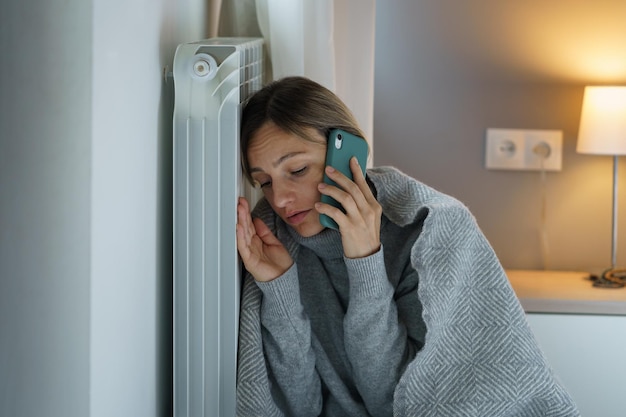 Mujer joven apoyada en el radiador de calefacción habla por teléfono con línea directa para resolver problemas con las facturas
