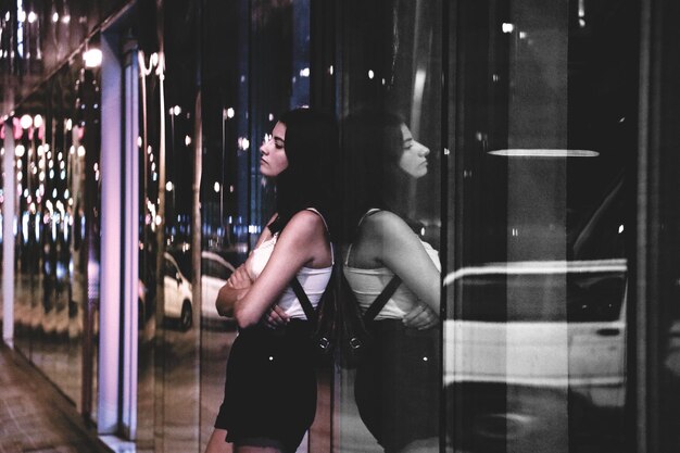 Foto mujer joven apoyándose en la pared de vidrio por la noche