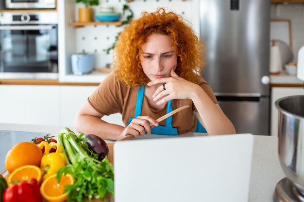 Una mujer joven aprende a cocinar, ve recetas en video en una computadora portátil en la cocina y cocina un plato Concepto de cocina en casa