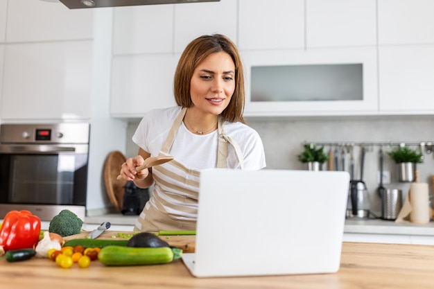 Una mujer joven aprende a cocinar, ve recetas de video en una computadora portátil en la cocina y cocina un plato.