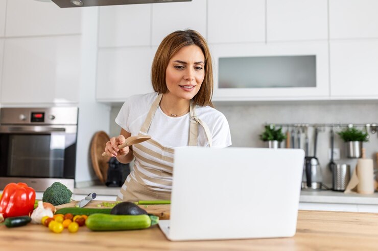 Foto una mujer joven aprende a cocinar, ve recetas de video en una computadora portátil en la cocina y cocina un plato.
