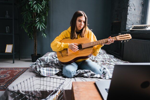 Mujer joven aprendiendo a tocar la guitarra en casa, viendo cursos en línea usando una computadora portátil, sentada en la cama