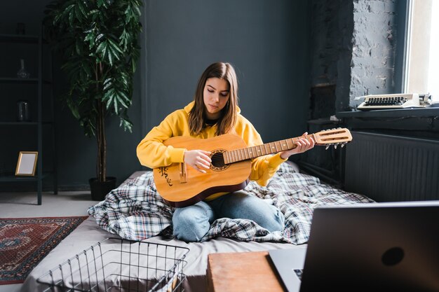 Mujer joven aprendiendo a tocar la guitarra en casa, viendo cursos en línea usando una computadora portátil, sentada en la cama