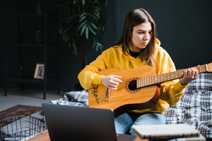 Foto mujer joven aprendiendo a tocar la guitarra en casa, viendo cursos en línea usando una computadora portátil, sentada en la cama