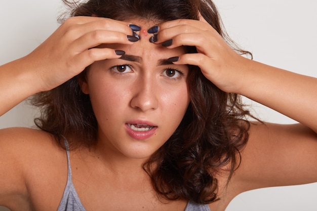 Mujer joven apretando grano en la frente, manteniendo la boca un poco abierta, con expresión facial triste