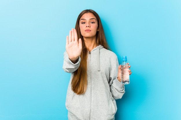 Mujer joven de la aptitud que sostiene una botella de agua que se coloca con la mano extendida que muestra la señal de stop, previniéndole.