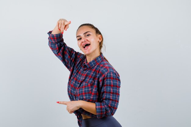 Mujer joven apuntando a la cámara y el lado izquierdo en camisa a cuadros, pantalones y mirando feliz, vista frontal.