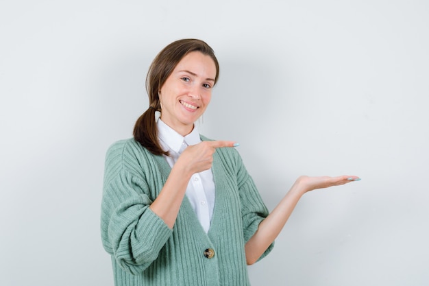 Mujer joven apuntando a su palma extendida a un lado en blusa, chaqueta de punto y mirando alegre, vista frontal.