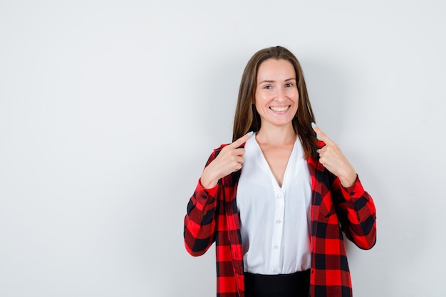 Mujer joven apuntando a su sonrisa en ropa casual y mirando alegre. vista frontal.