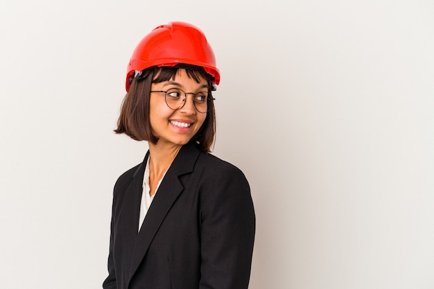 Mujer joven arquitecto con casco rojo aislado sobre fondo blanco mira a un lado sonriente, alegre y agradable.