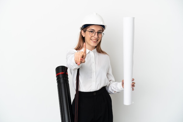 Mujer joven arquitecto con casco y sosteniendo planos aislados en blanco mostrando y levantando un dedo