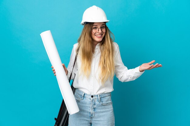 Mujer joven arquitecto con casco y sosteniendo planos aislados en espacio azul con expresión facial sorprendida