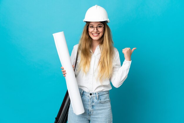 Mujer joven arquitecto con casco y sosteniendo planos aislados en la pared azul apuntando hacia el lado para presentar un producto