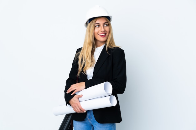 Mujer joven arquitecto con casco y sosteniendo planos sobre blanco aislado mirando hacia el lado