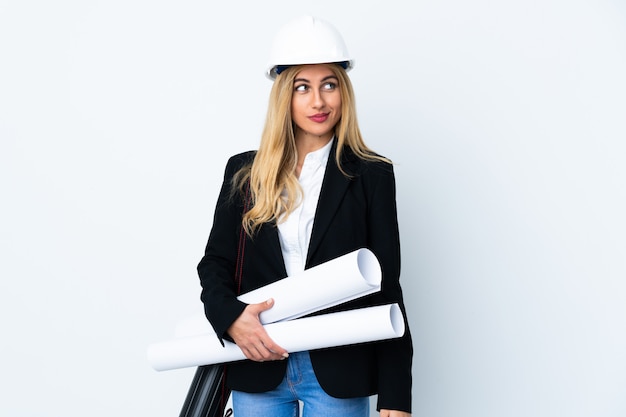 Mujer joven arquitecto con casco y sosteniendo planos sobre espacio en blanco aislado de pie y mirando hacia el lado