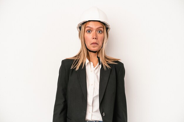 Mujer joven arquitecto caucásico con un casco aislado sobre fondo blanco se encoge de hombros y abre los ojos confundidos.