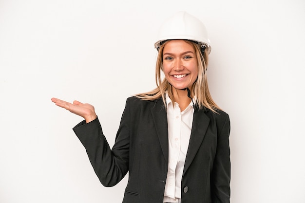 Mujer joven arquitecto caucásico con un casco aislado sobre fondo blanco que muestra un espacio de copia en una palma y sosteniendo otra mano en la cintura.