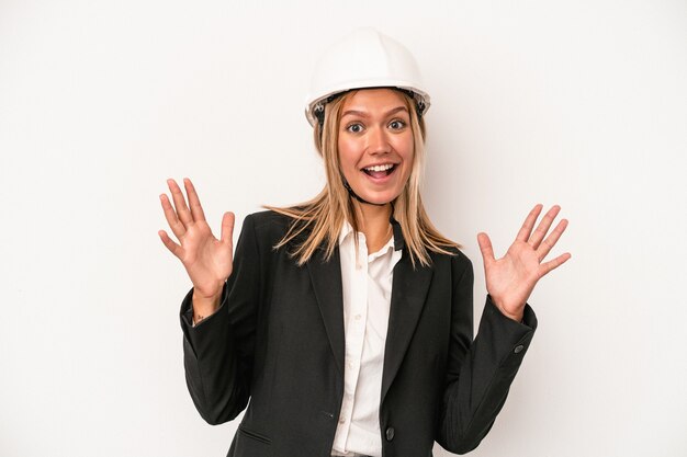 Mujer joven arquitecto caucásico con casco aislado sobre fondo blanco recibiendo una agradable sorpresa, emocionada y levantando las manos.