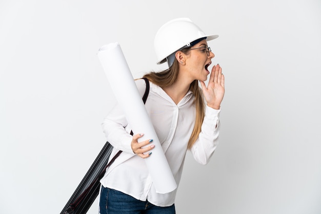 Mujer joven arquitecto lituano con casco y sosteniendo planos aislados sobre fondo blanco gritando con la boca abierta hacia el lado