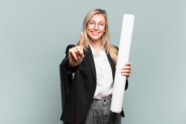 Mujer joven arquitecto sonriendo y mirando amistosamente, mostrando el número uno o el primero con la mano hacia adelante, contando hacia atrás