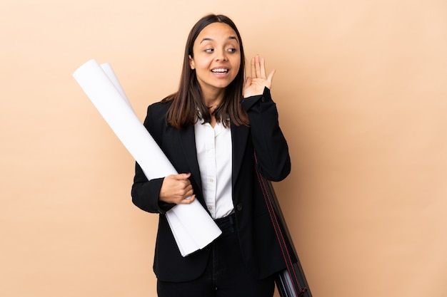 Mujer joven arquitecto sosteniendo planos sobre pared aislada escuchando algo poniendo la mano en la oreja