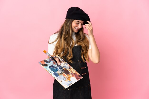 Mujer joven artista sosteniendo una paleta aislada en la pared rosa riendo