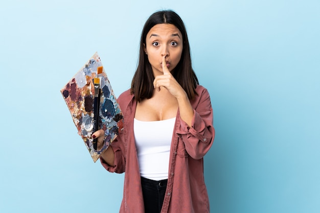 Mujer joven artista sosteniendo una paleta sobre azul aislado mostrando un signo de silencio gesto poniendo el dedo en la boca.