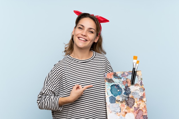 Mujer joven artista sosteniendo una paleta sobre fondo azul aislado y apuntando