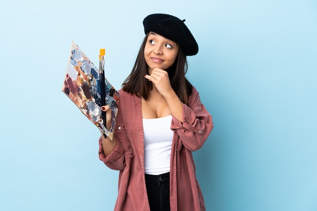 Mujer joven artista sosteniendo una paleta sobre fondo azul aislado pensando en una idea mientras mira hacia arriba.