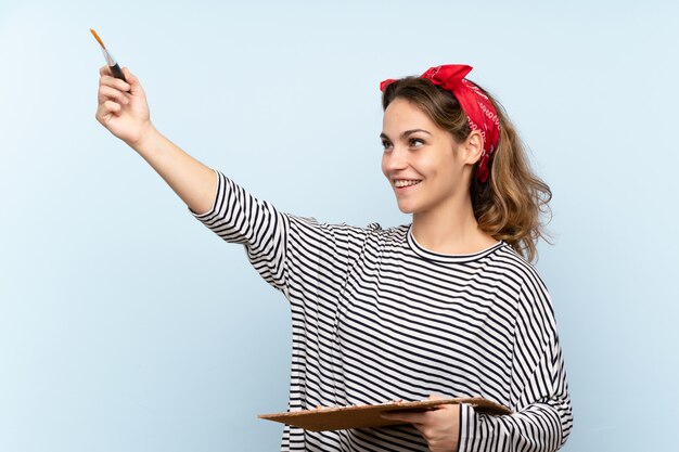 Mujer joven artista sosteniendo una paleta sobre pared azul aislada