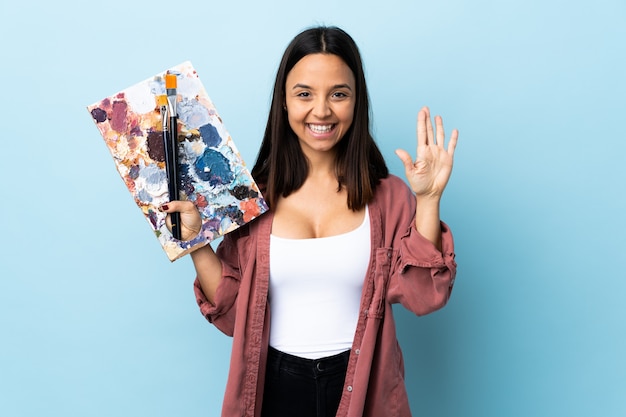 Mujer joven artista sosteniendo una paleta sobre pared azul saludando con la mano con expresión feliz.