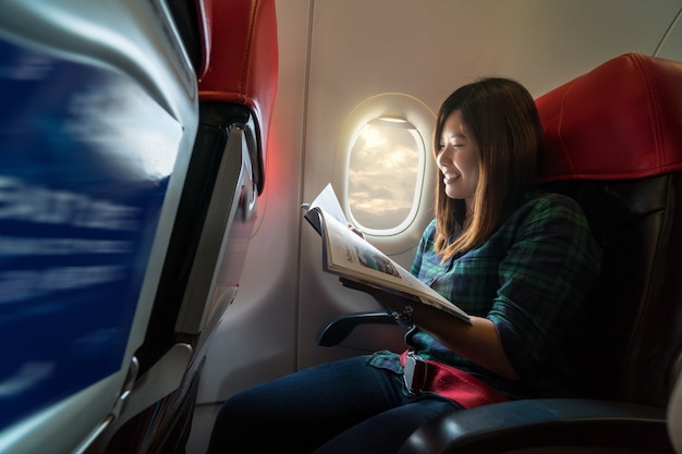 Mujer joven de Asia leyendo la revista mientras viaja dentro del avión al lado del windo