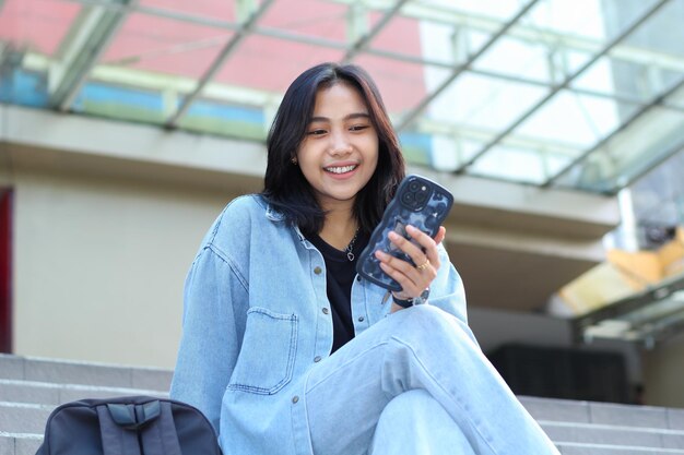 mujer joven asiática elegante y sonriente sin preocupaciones usando una aplicación de teléfono inteligente video chat tienda en línea y navegando por las redes sociales mientras está sentada en las escaleras de un centro comercial ropa casual al aire libre