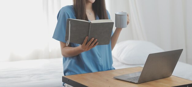 Mujer joven asiática con hermosa cara, cabello largo y camisa azul acostada en la cama en un dormitorio blanco en casa con una taza de café y una llamada de video con una computadora portátil hablando con parientes en vacaciones concepto de vacaciones
