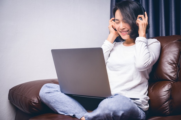 Mujer joven asiática hermosa que se relaja con su música que juega con el ordenador portátil en el sofá. Trabajo en casa y concepto de la felicidad.