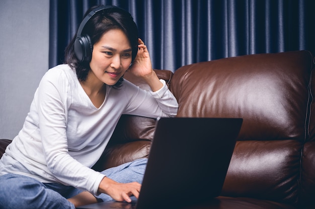 Mujer joven asiática hermosa que se relaja con su música que juega con el ordenador portátil en el sofá. Trabajo en casa y concepto de la felicidad.