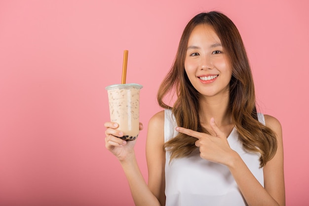 Mujer joven asiática hermosa sosteniendo beber té de leche de burbuja de perla de tapioca con sabor a azúcar moreno y señalando con el dedo, retrato de sonrisa mujer, aislada en fondo rosa, concepto de bebida de té de leche de perla