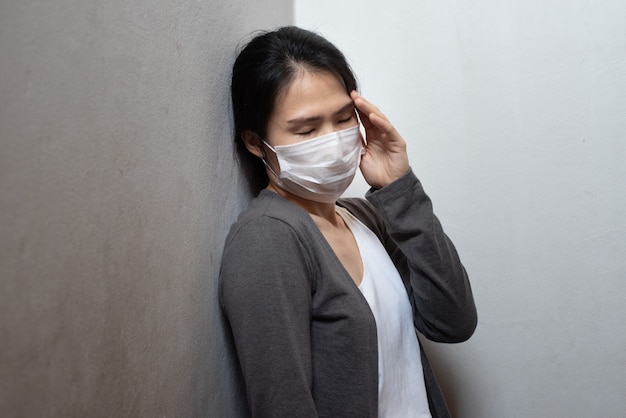 Mujer joven asiática con máscara quirúrgica sintiendo dolor de cabeza enfermo y tos apoyándose en la pared aislada sobre fondo blanco. Prevención de brotes de coronavirus de Wuhan (COVID-19). Concepto de salud