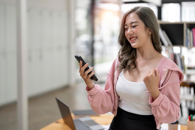 Mujer joven asiática mirando el teléfono móvil riendo con buenas noticias Mujer feliz y alegre mirando la aplicación del teléfono celular leer el mensaje sentirse emocionada